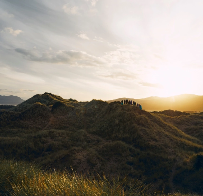 A group of people walking over sand dunes with the sun setting in the background
