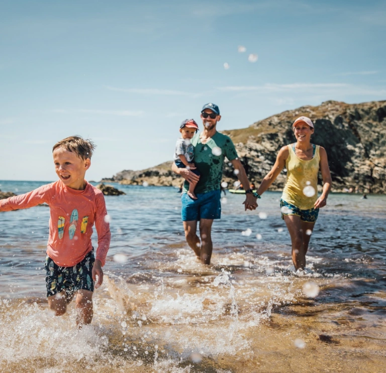 Family of two children and two parents splashing about in the shallow sea water on a beach in the sunshine. There are rocks near the sea in the distance.