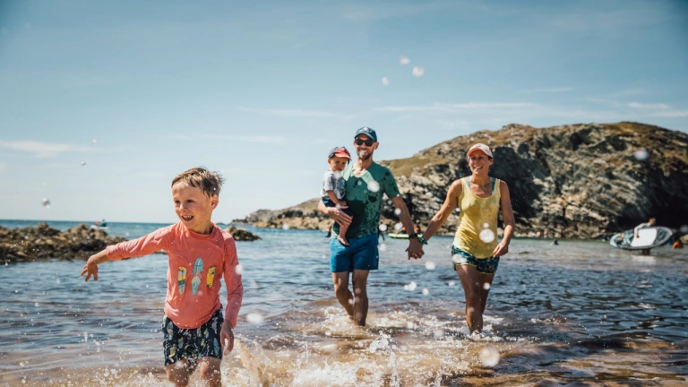 Family of two children and two parents splashing about in the shallow sea water on a beach in the sunshine. There are rocks near the sea in the distance.