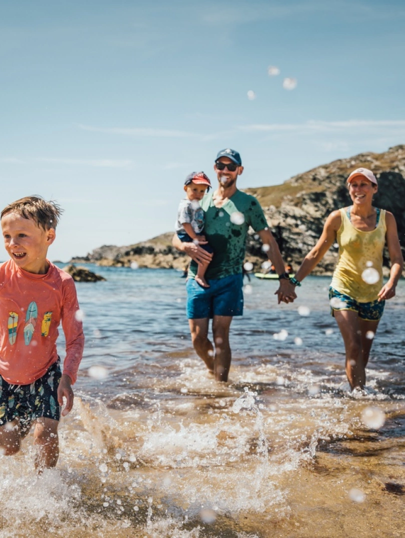 Family of two children and two parents splashing about in the shallow sea water on a beach in the sunshine. There are rocks near the sea in the distance.