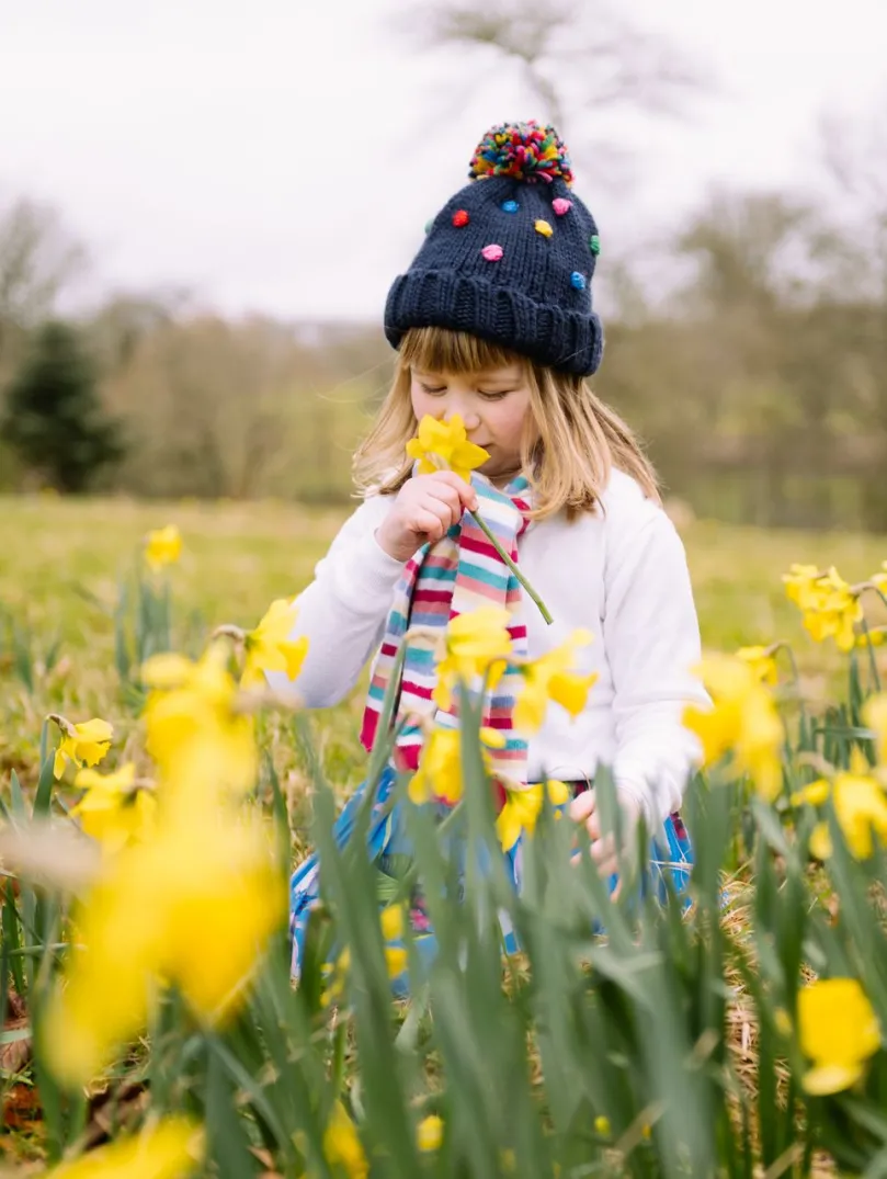 A young girl stands in a field of bright yellow daffodils in spring. She wears a navy knitted hat with a pom-pom, a striped scarf, and a white jumper, sniffing a flower. 