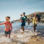 Family of two children and two parents splashing about in the shallow sea water on a beach in the sunshine. There are rocks near the sea in the distance.